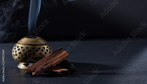 Pieces of dark oud wood beside a pierced brass incense burner with rising smoke on dark stone, Arabian bakhoor still life with generous copy space