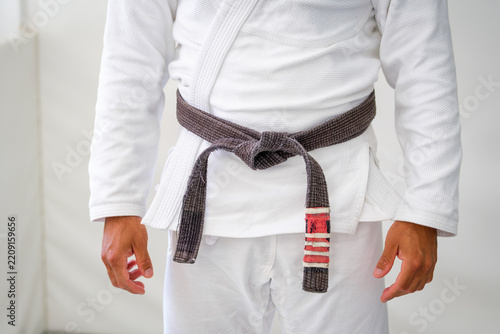 Close up of a worn Brazilian jiu jitsu brown belt tied over a white gi, with rank stripes on one end and the hands of the practitioner at the sides