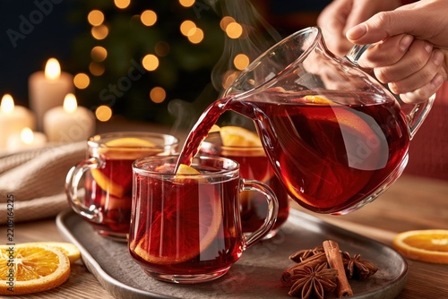 Hot mulled wine being poured from glass pitcher into clear mugs with orange slices, cinnamon sticks and star anise on wooden tray, warm candlelight background