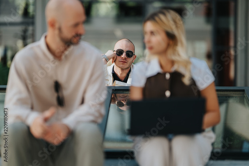 Man in sunglasses leans on a railing while looking toward two people sitting nearby. A blurred couple uses a laptop on outdoor steps with an onlooker in the background.