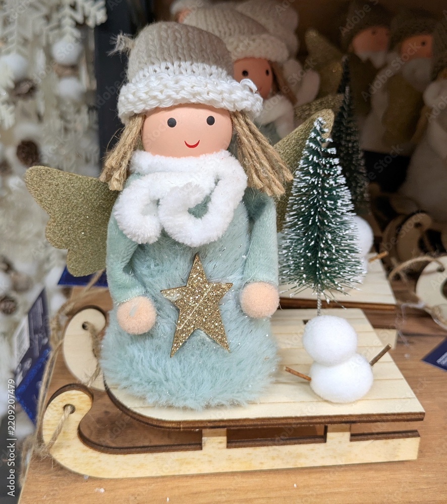 Fototapeta premium Decoración de Navidad. Muñeco de un ángel con alas doradas, con un sombrero de lana, bufanda y túnica con una estrella, colocado sobre un trineo de madera con un árbol de navidad