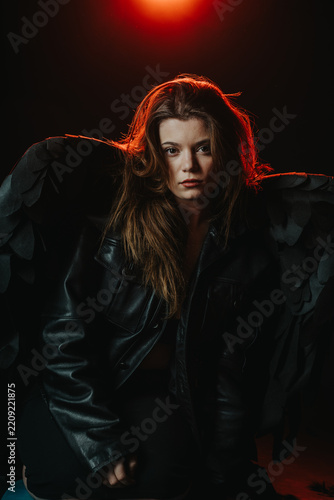 A young woman posed wearing black wings and a leather jacket in dramatic lighting. Moody studio portrait with red backlight highlighting her hair and dark costume.