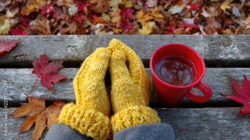 A person wearing yellow knitted gloves holds a coffee mug in their hands, sitting on an old wooden bench surrounded by autumn leaves