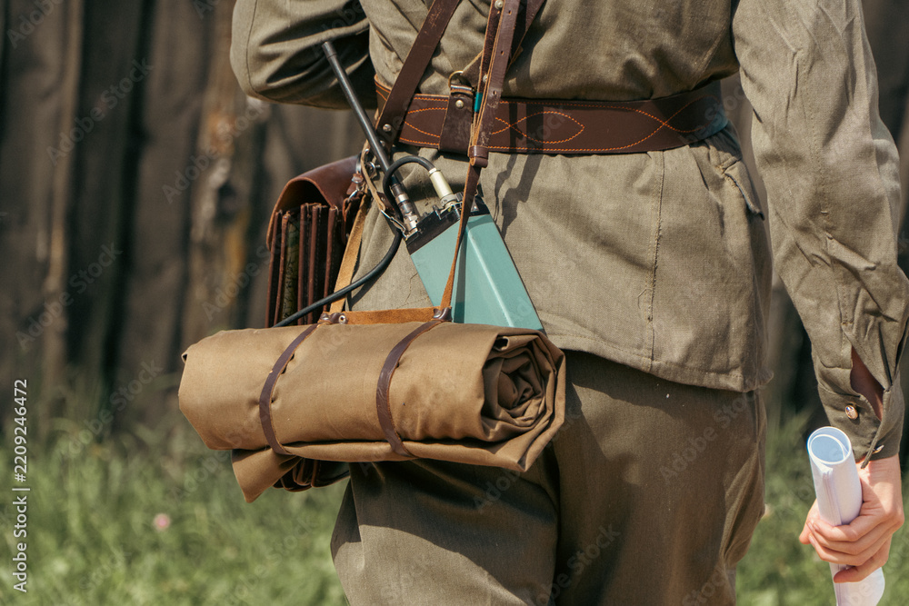 © Dmitry - A person wearing a vintage military uniform carries a portable radio transceiver, a brown leather map case, and a rolled canvas mat © Dmitry - A person wearing a vintage military uniform carries a portable radio transceiver, a brown leather map case, and a rolled canvas mat