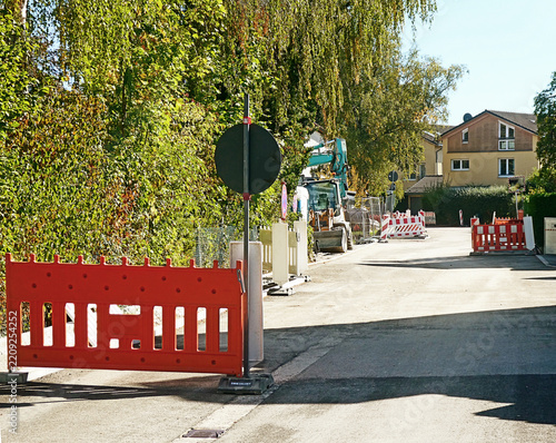 Road partially blocked with red and white barrage,  in view a bulldozer with shovel in pause and contruction material
