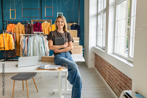 A young woman stands confidently in her small clothing business, holding a tablet. The store features colorful garments displayed on racks and boxes stacked nearby. Natural light floods the space, cre