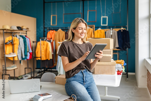 A young woman smiles while using a tablet to manage her small clothing business. She sits on a table surrounded by cardboard boxes and colorful clothing displays in a bright, modern boutique.