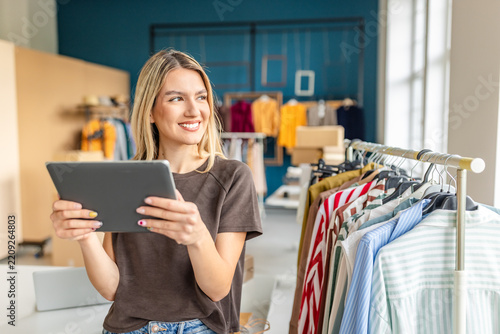 A young woman smiles while holding a tablet in her small clothing store. The shop features a variety of colorful garments displayed on racks, creating a vibrant and inviting atmosphere.