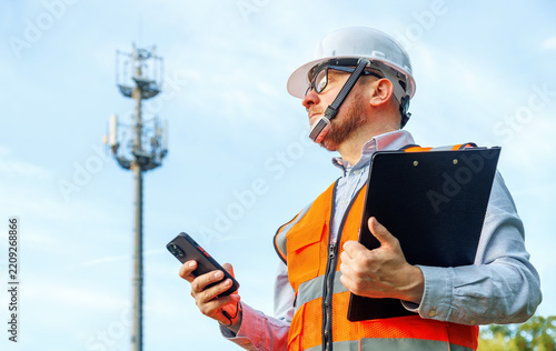 Telecom engineer using AI powered mobile app on smartphone near 5G base station. Technician in hardhat with clipboard running AI network diagnostics, signal audit and site survey.