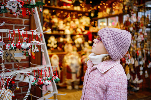 Little school girl on Christmas market. Child in a shop with Christmas tree decorations and Santa Clause figures.