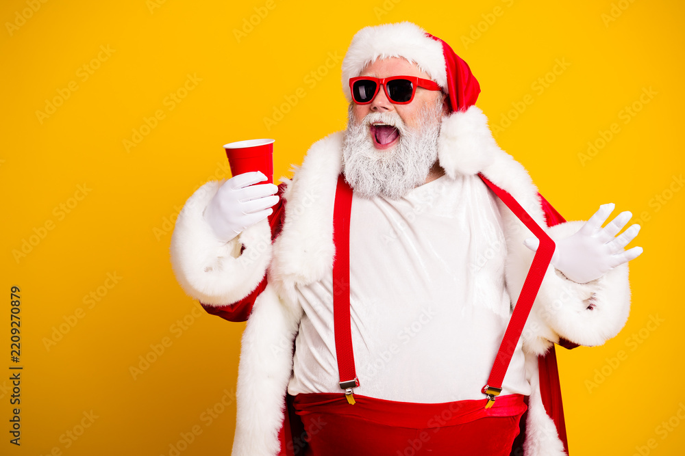 Obraz premium Cheerful Santa Claus in sunglasses holding a red party cup celebrating Christmas and holiday fun in a festive red and white outfit against a bright yellow background