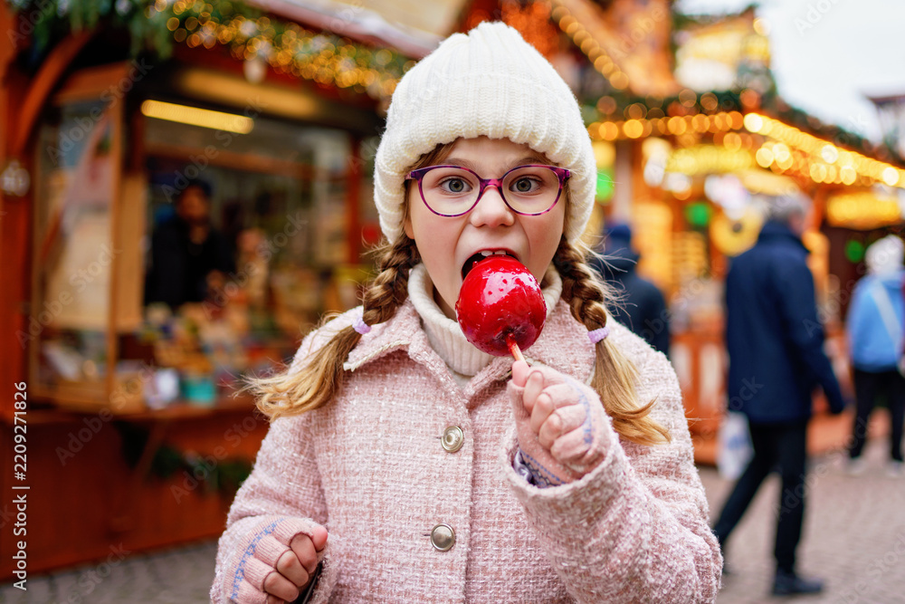 Obraz premium Little cute kid girl eat sweet sugar covered red candy apple on Christmas market. Happy child on traditional Xmas fair in Germany.