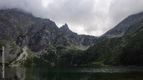 View of Morskie Oko lake surrounded by rocky peaks and lush greenery under a cloudy sky in Tatra County, Lesser Poland Voivode, Poland.