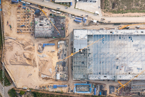 Oblique aerial view of an expanding factory construction project with a broad steel roof deck, cranes, foundations and site roads at Eastern Seaboard Industrial Estate, Rayong, Thailand.