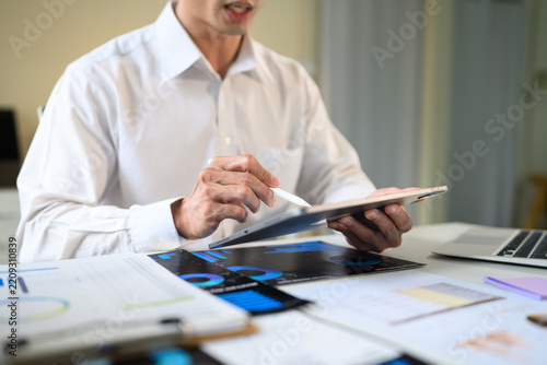 Businessman Reviewing Data Charts on Tablet with Stylus at Office Desk Surrounded by Printed Reports