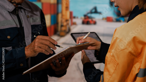 Warehouse workers review a clipboard and tablet while checking inventory in a busy loading area with containers and forklift equipment.
