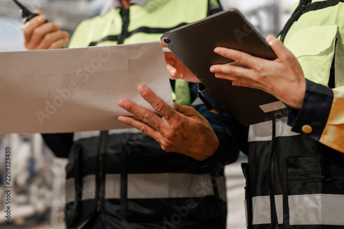 Close-up of construction workers in safety vests reviewing a blueprint and tablet at a work site, teamwork, planning and project management.