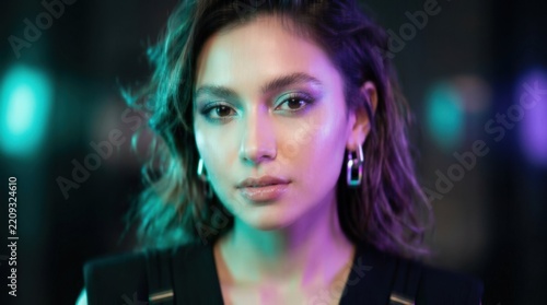Close frontal beauty portrait illuminated by turquoise-green and violet light, face softened by motion and shallow focus, dark neutral background, luminous skin and futuristic high-fashion cosmetics