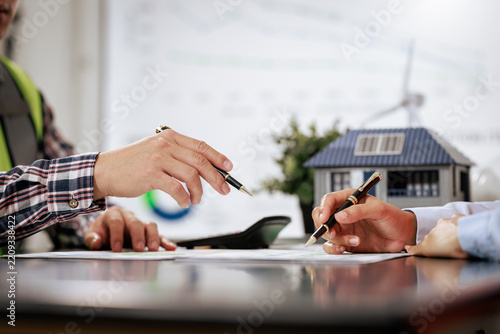 People signing property documents beside a model house with solar panels, a home purchase, real estate agreement, and renewable energy planning.