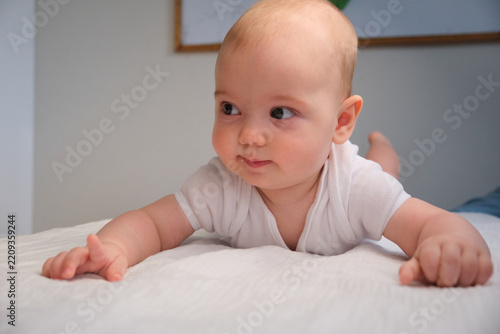 Cute baby lies on white bed and looks around with curious facial expression