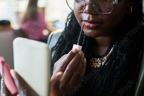 Close up shot of young adult African American woman applying lip gloss while looking at smartphone in creative office. Scene suitable for beauty campaigns and cosmetics marketing