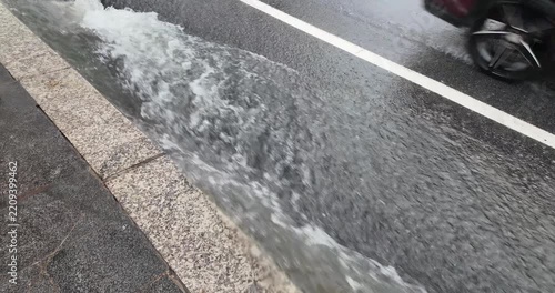 Slow motion capture of a torrent of rainwater rushing along the sidewalk during a downpour, while vehicles drive by nearby.