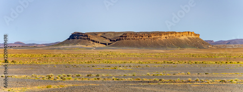 A wide view shows a rock formation in the dry desert of Morocco. The area is open with sparse vegetation and clear skies. The rock stands alone in the vast land.