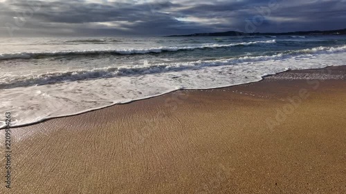 waves roll onto a sandy beach against a background of blue sky with dramatic thunderclouds, sunset, beautiful landscape, slow motion