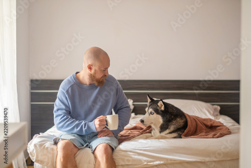 Obraz na plátně Man looks at his husky holding white cup of coffee on wide bed, together without needing any words, moment building real trust