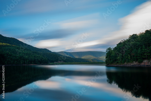 Long exposure landscape of Switzer Lake in Virginia at dusk, capturing smooth, glass-like water and softly blurred clouds passing over dark forested mountains during a moody twilight.