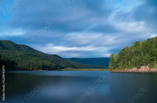 Long exposure landscape of Switzer Lake, Virginia, showing a mountain reservoir with smooth water reflecting a cloudy sky. Lush green forests cover the surrounding hills in this tranquil wilderness.