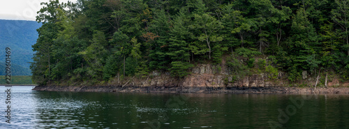 Close-up view of the rugged, rocky shoreline of Switzer Lake, Virginia, topped with dense green pine and deciduous trees meeting the calm waters in a pristine, untouched wilderness setting.