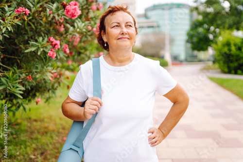 Older woman stands on a park path with a yoga mat over her shoulder. Mature woman in sportswear is preparing for a workout on a sunny day. Concepts: sports, healthy lifestyle, relaxation.