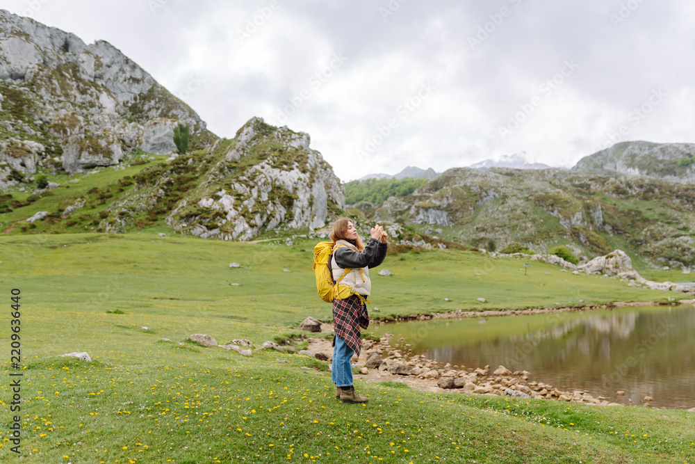 Fototapeta premium Female hiker with a yellow backpack uses her phone to photograph a mountain-ringed lake. She is enjoying nature and holding her smartphone on a cloudy day. Nature and adventure concept.