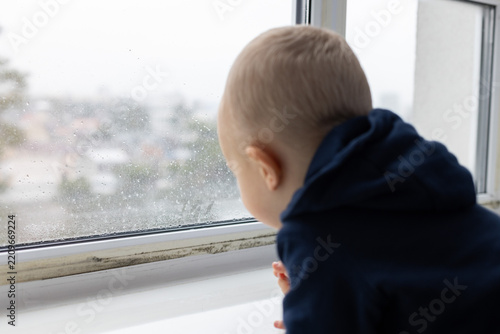 Little child looking through the window with water drops condensate and black spots of mouldy fungus on it. Selective focus.