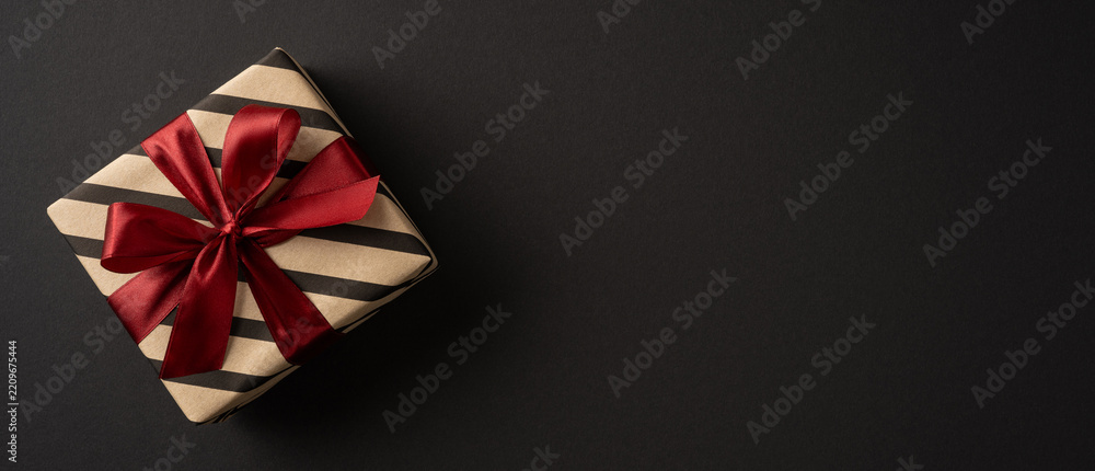 Obraz premium Wrapped gift with red satin bow on banner black wide background for Merry Christmas and Happy New Year