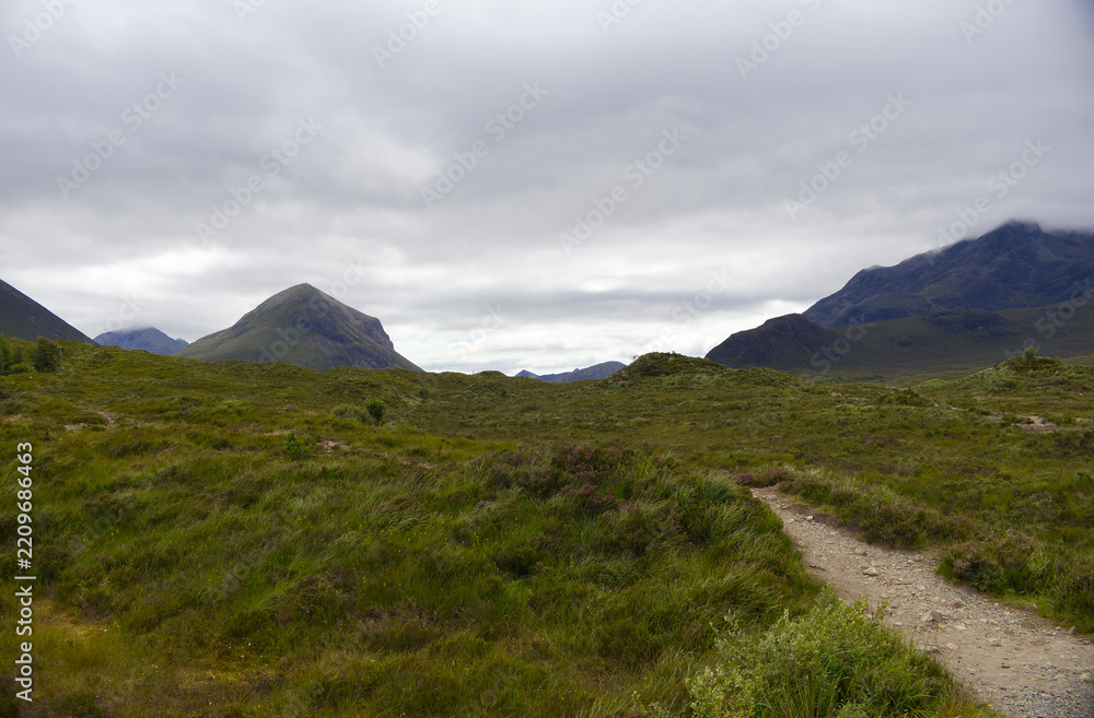 Fototapeta premium Hiking trail leading through green moorland towards the dramatic dark peaks of the Black Cuillin mountains on the Isle of Skye, Scotland.