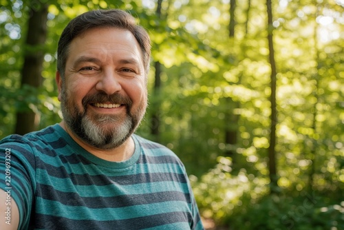 Cheerful plump middle aged man smiles for selfie in sunny park. Bearded caucasian male in striped t shirt enjoys summer nature walk. Happy overweight guy feels confident, relaxed spending time