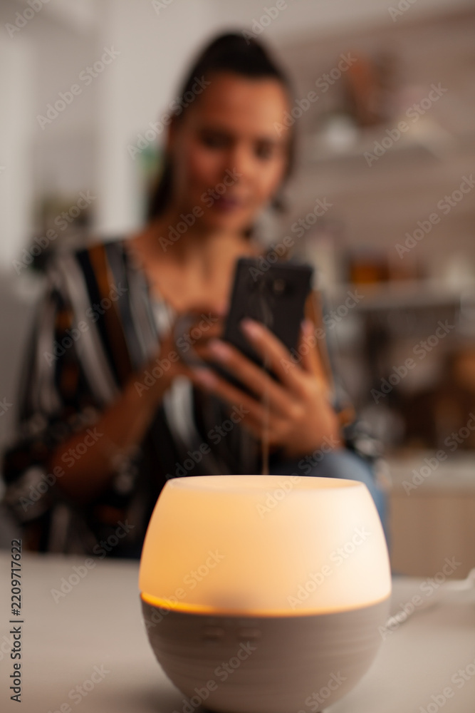 Fototapeta premium Essential oil diffuser glows in sharp foreground focus while a woman uses her smartphone and holds a mug in the background, suggesting calm digital downtime at home, supporting aromatherapy use.
