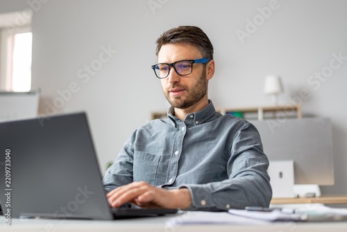 Focused Caucasian Man In His 30s Working On Laptop At A Modern Office Desk, Engaged In Professional Tasks. Creating, Typing, Or Reviewing Documents While Surrounded By Office Equipment.