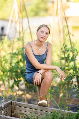 Rural dweller girl in sundress with garden tools against background of vegetable garden, resting between jobs, gardening, growing plants on house plot