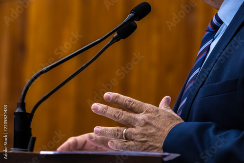Close-up of a businessman or politician wearing a green suit and watch gesturing during a speech with microphones at a podium. 