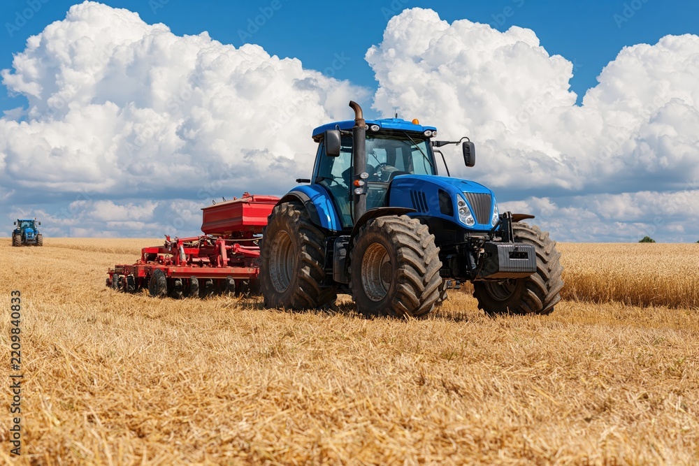 Obraz premium Blue tractor with seed drill working in golden wheat field under cloudy summer sky