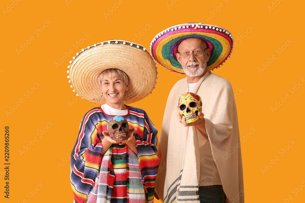 Obraz premium Dia de los muertos celebration, happy senior couple wearing traditional mexican sombreros and ponchos holding decorated sugar skulls on orange background