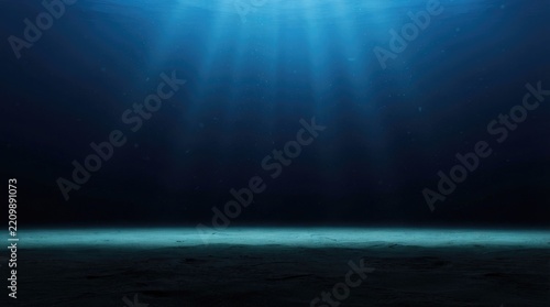 Sunbeams penetrate deep into a dark, vast underwater environment. A horizontal sandy seabed stretches across the bottom of the image, illuminated by the light above.