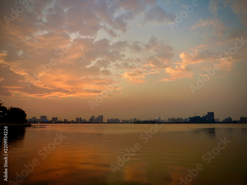 Golden Sunset Reflections Across a Peaceful Urban Lake with a Distant City Skyline, Warm Orange Clouds and Calm Evening Water on West Lake in Hanoi, Vietnam