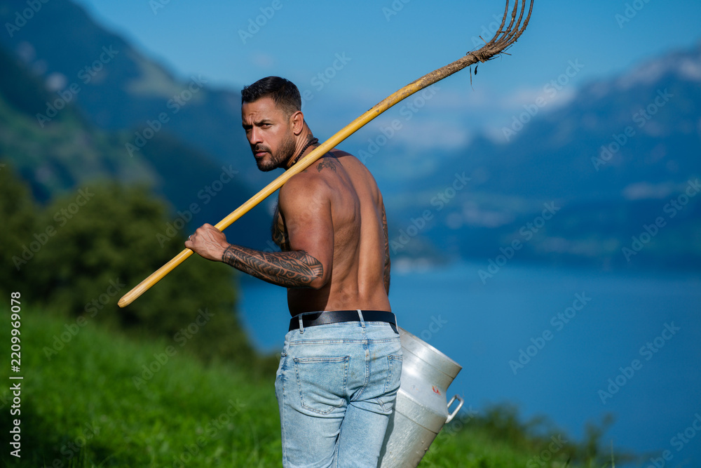 Obraz premium Eco farm portrait with muscular dairy farmer. Traditional eco farm lifestyle with rustic pitchfork. Eco farm worker carrying milk can outdoors. Shirtless man in eco farm dairy portrait.