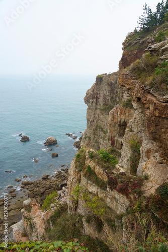 Rugged cliff rising from the sea on the northeastern China coast near Dalian, with rocky shoreline, green vegetation and calm blue water under a hazy sky