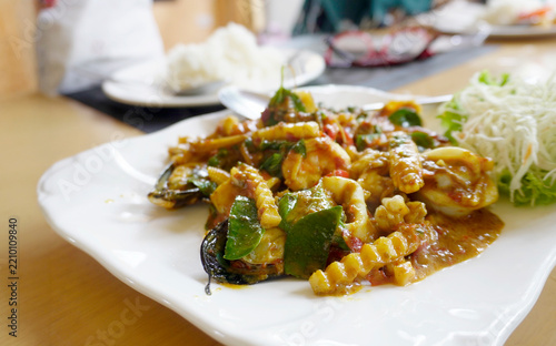 elicious plate of spicy Thai stir-fried seafood with squid, mussels, and herbs on a white plate, served on a table in a restaurant.