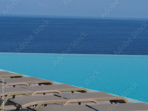View of the infinity pool with sun loungers against the backdrop of the sea, and the hotel grounds.